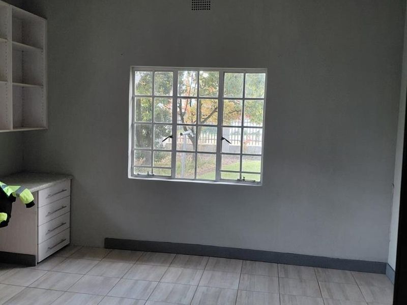 A clean, empty bedroom featuring neutral grey walls, light-toned tiled flooring, and built-in desk and shelving units.