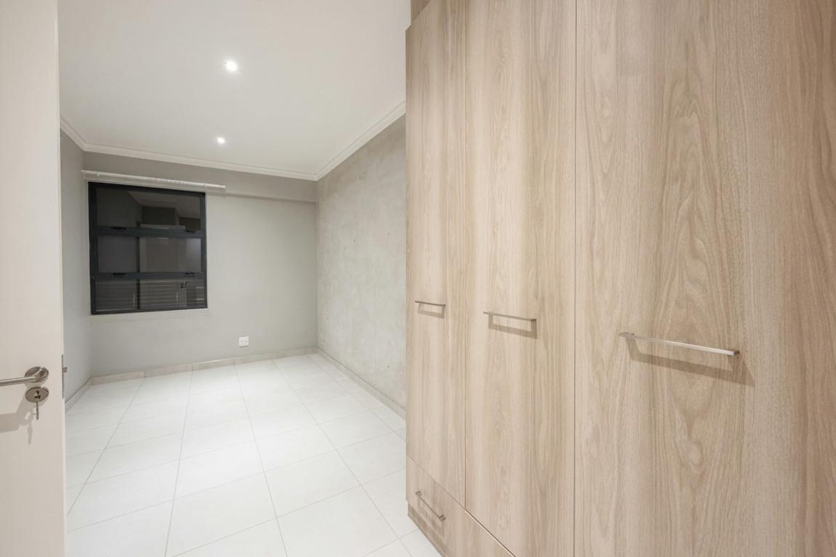 A bright, empty bedroom featuring modern light wood-finish built-in cupboards, neutral tiled flooring, and recessed downlighting.