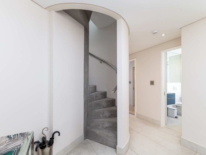 A bright, modern hallway featuring a distinctive curved staircase with a metal handrail, tiled flooring, and clean, neutral finishes.