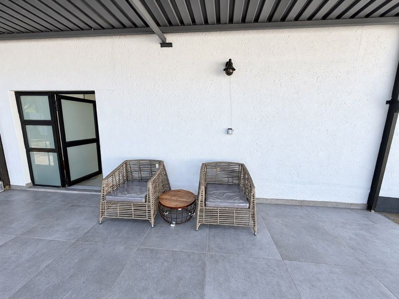 A covered outdoor patio area featuring large format grey floor tiles, a modern wall-mounted light, and a sleek metal roof structure.
