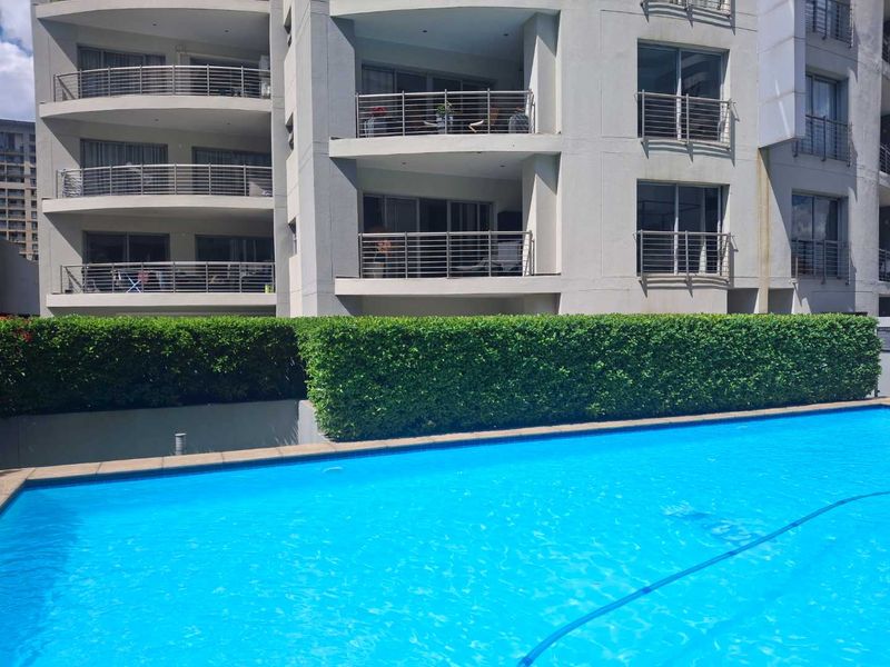 A view of the apartment complex swimming pool area featuring landscaped hedges and multiple balconies with stainless steel railings.