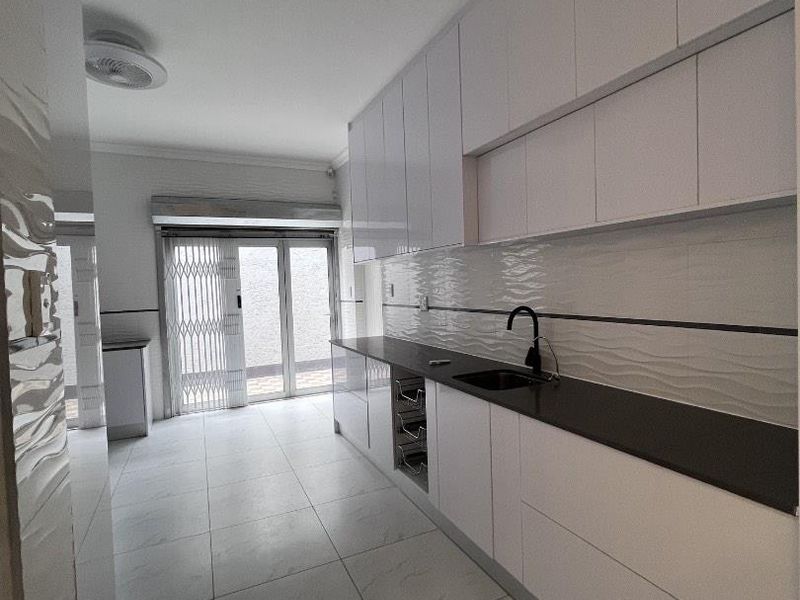 Modern galley-style kitchen featuring sleek white cabinetry, dark stone countertops, textured backsplash tiles, and large format floor tiles with direct access to the patio.
