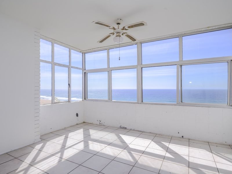 An empty, sunlit enclosed balcony featuring tiled floors, painted brick walls, and expansive ocean views through large windows.