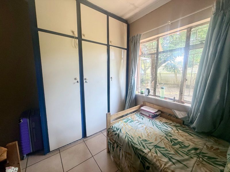 A compact bedroom featuring built-in cupboards, tiled flooring, and a steel-framed window with security bars.