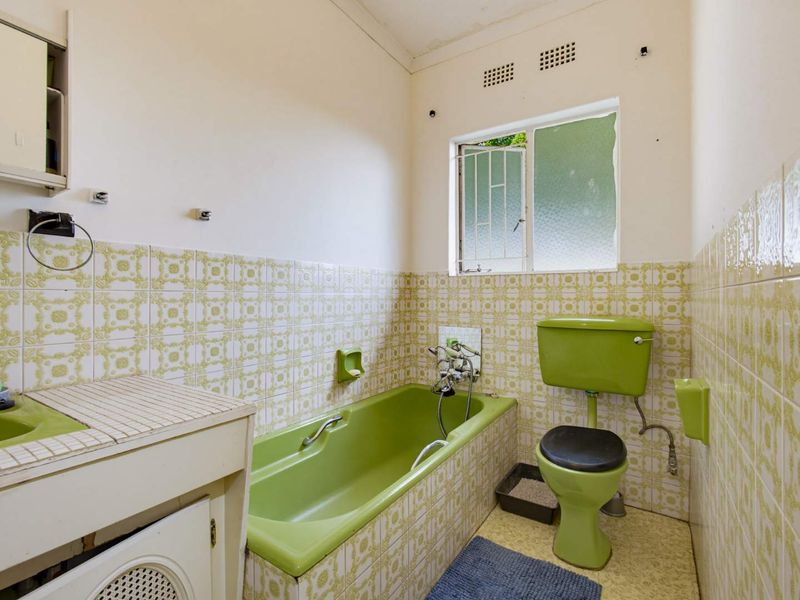 A dated bathroom featuring original green sanitary ware, patterned wall tiles, and a window with security bars, requiring modernization.