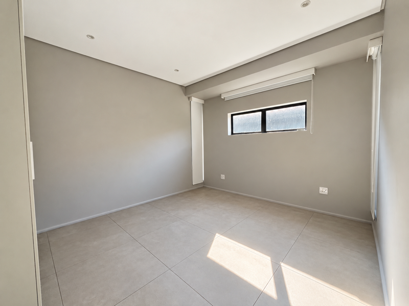 A bright, empty bedroom featuring neutral grey walls, large format floor tiles, recessed lighting, and built-in storage with roller blinds.