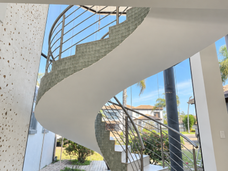 A bright, modern entrance hall featuring a striking spiral staircase with stainless steel railings and expansive floor-to-ceiling windows overlooking the garden.
