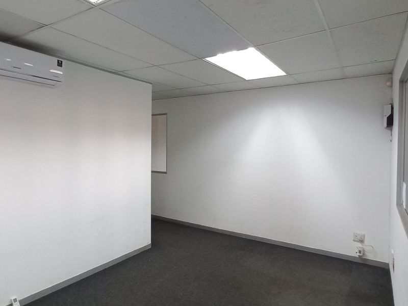 A clean, empty office space featuring a suspended ceiling, recessed lighting, dark carpeting, and a wall-mounted air conditioning unit.