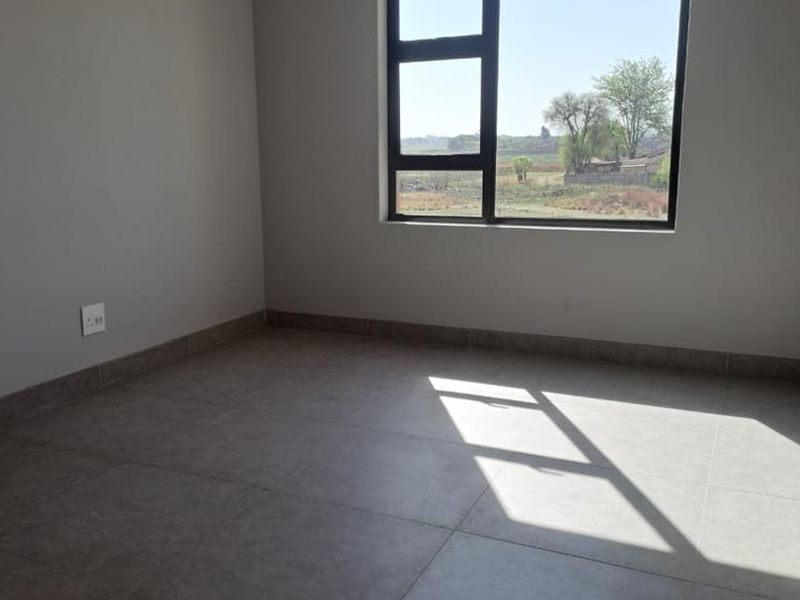 A bright, empty bedroom featuring neutral grey walls, large aluminium-framed windows, and modern tiled flooring, offering a clean and versatile living space.
