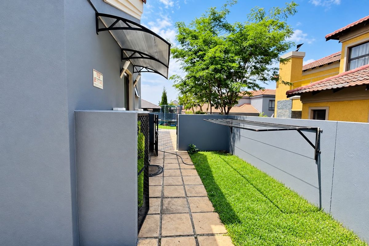 A well-maintained rear exterior area featuring a paved walkway, a neat lawn, a wall-mounted drying rack, and a protective polycarbonate awning over the entrance.