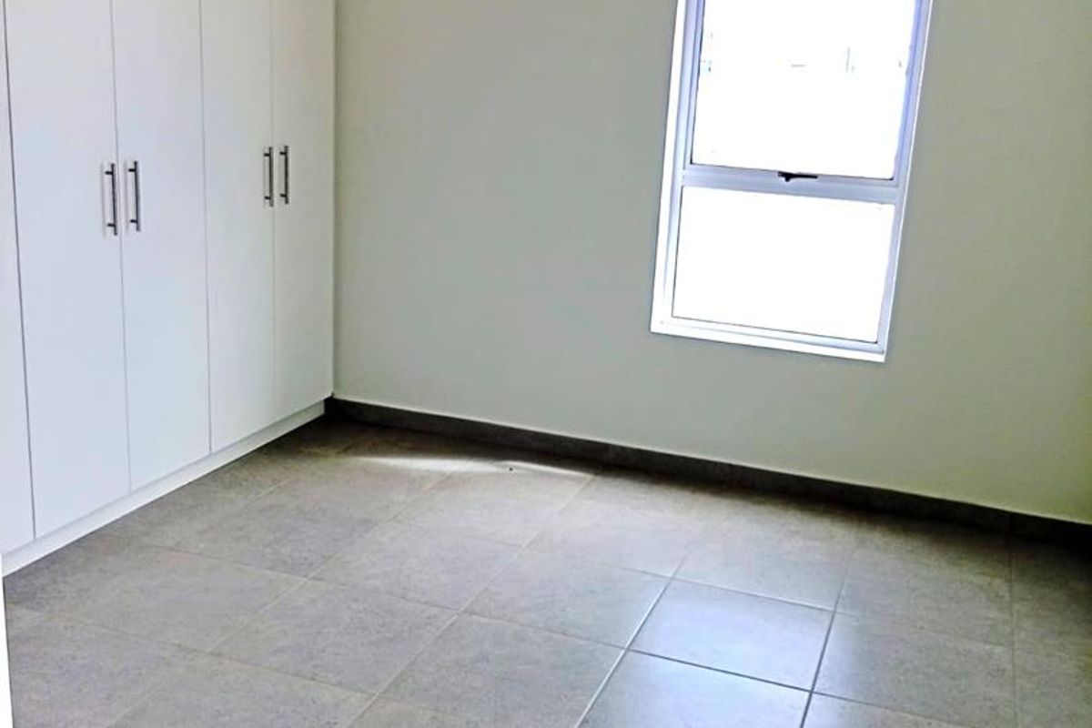 A clean, empty bedroom featuring neutral tiled flooring, built-in white cupboards, and a single window providing natural light.