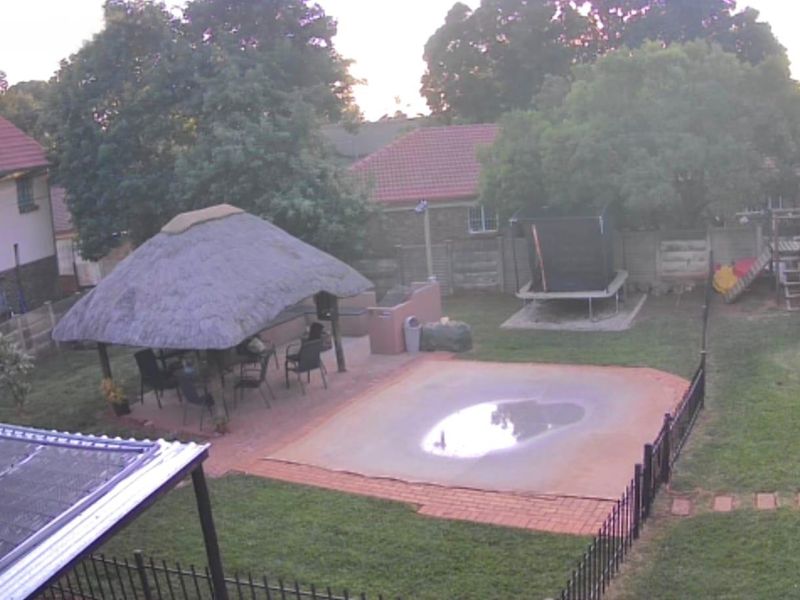 Rear garden view featuring a thatched lapa, swimming pool with cover, brick paved patio area, and perimeter walling.