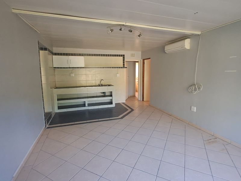 An open-plan living area featuring tiled flooring, a basic built-in kitchenette with sink, and a wall-mounted air conditioning unit.