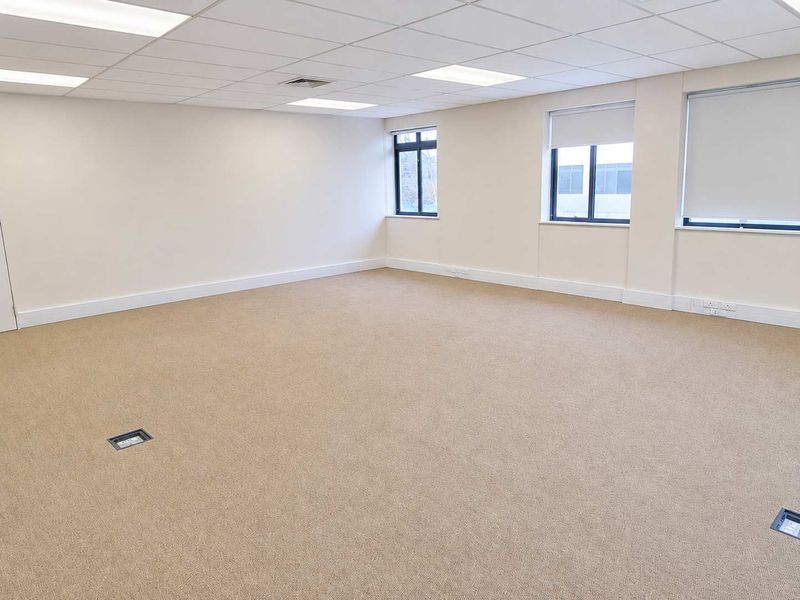 A spacious, empty office area featuring neutral carpeting, suspended ceiling with recessed lighting, and large windows with roller blinds.