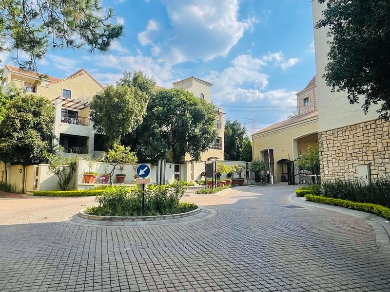 A well-maintained apartment complex entrance featuring a paved circular driveway, stone facade detailing, and established landscaped gardens.