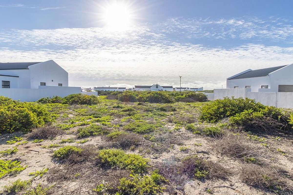 A vacant residential plot featuring natural coastal vegetation, surrounded by established white-walled residential properties in a quiet estate setting.