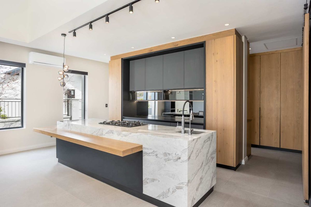 Modern kitchen featuring a large marble island with integrated gas cooktop, sleek dark cabinetry, wood-toned accents, and contemporary track lighting.
