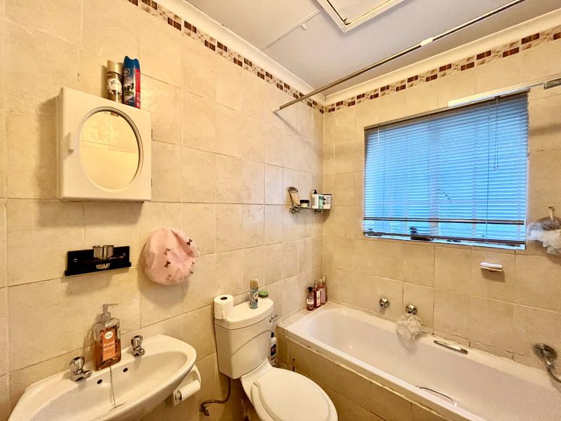 A functional bathroom featuring a built-in bathtub, pedestal basin, and toilet, finished with neutral ceramic wall tiles and a window for natural light.