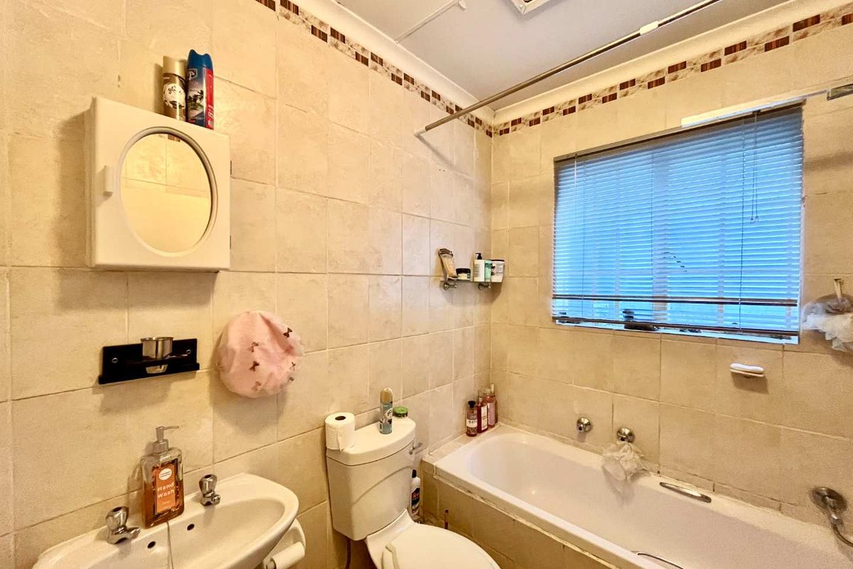 A functional bathroom featuring a built-in bathtub, pedestal basin, and toilet, finished with neutral ceramic wall tiles and a window for natural light.
