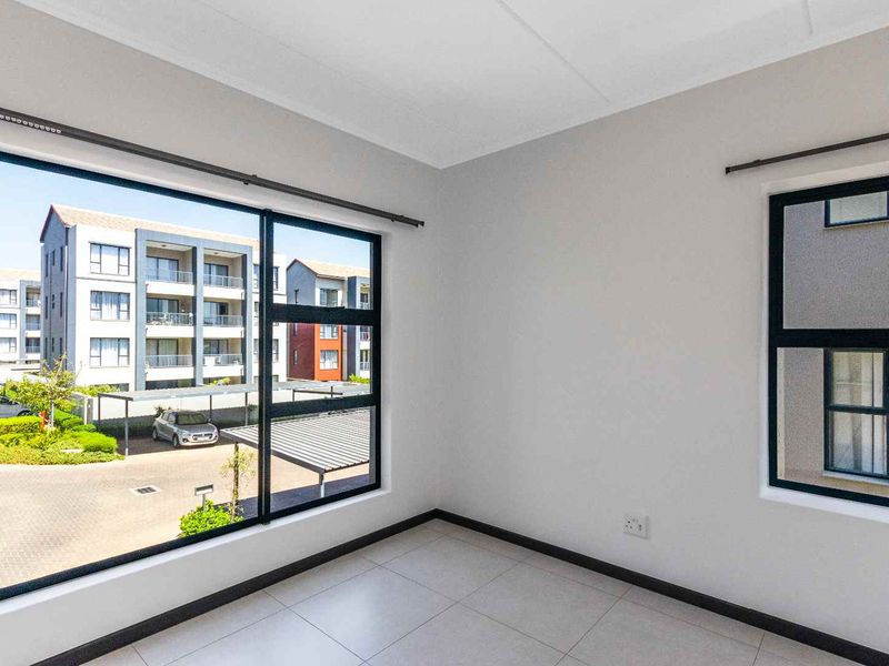 A bright, empty bedroom featuring neutral tiled flooring, modern aluminium window frames, and ample natural light with views of the surrounding apartment complex.
