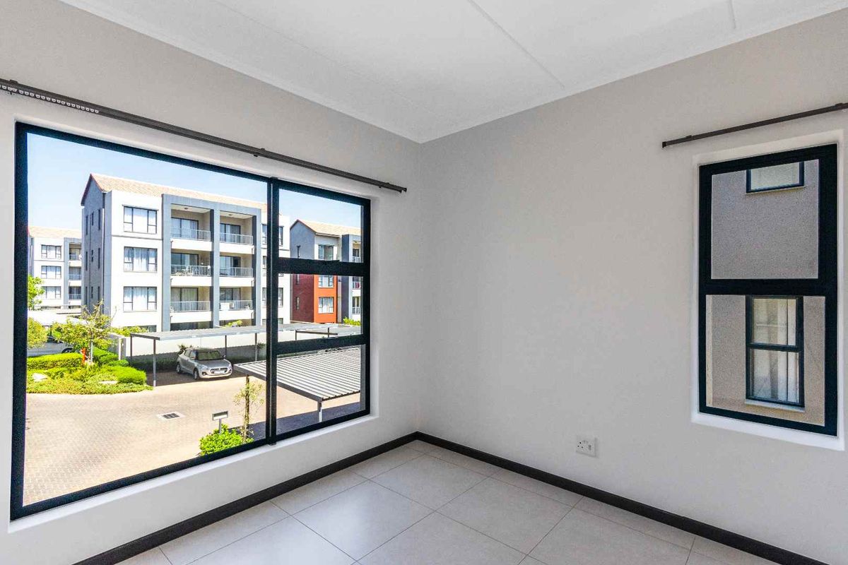 A bright, empty bedroom featuring neutral tiled flooring, modern aluminium window frames, and ample natural light with views of the surrounding apartment complex.