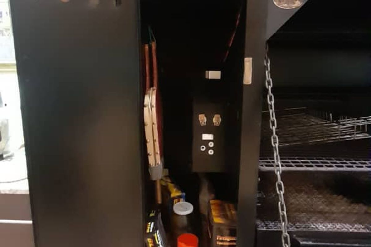 A built-in indoor braai or fireplace unit featuring a black metal finish, integrated shelving, and a chain-operated grill mechanism.