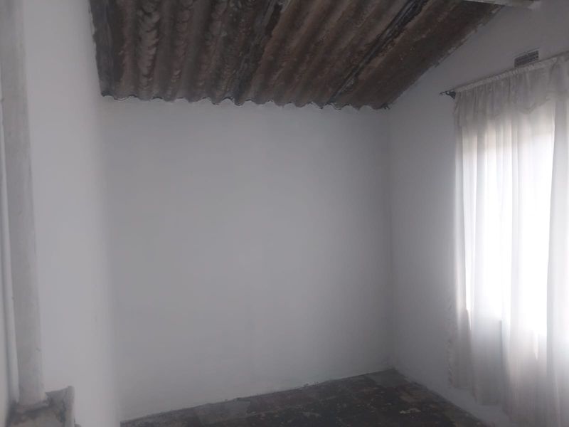 An empty bedroom featuring a corrugated iron ceiling and a window with a curtain rail, requiring significant renovation to improve finishes.