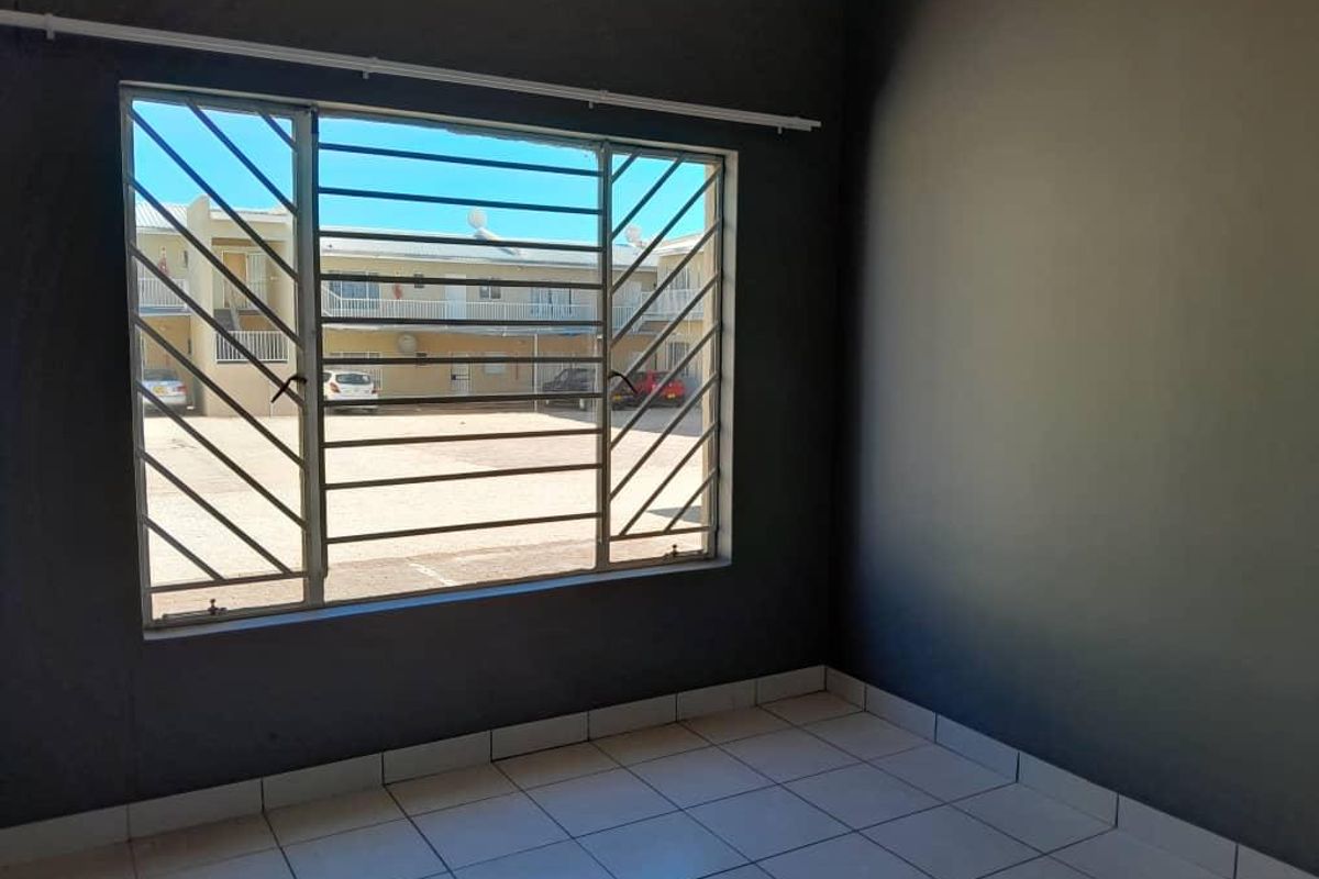 An empty bedroom featuring tiled flooring, dark grey walls, and a window fitted with security bars overlooking the apartment complex courtyard.