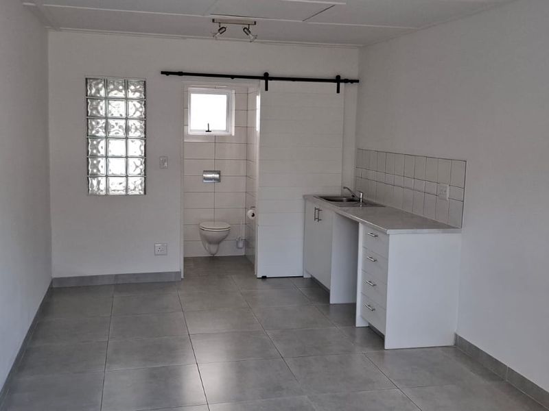 An open-plan studio space featuring tiled flooring, a compact kitchenette with sink, and an integrated bathroom with a wall-mounted toilet and glass block window.