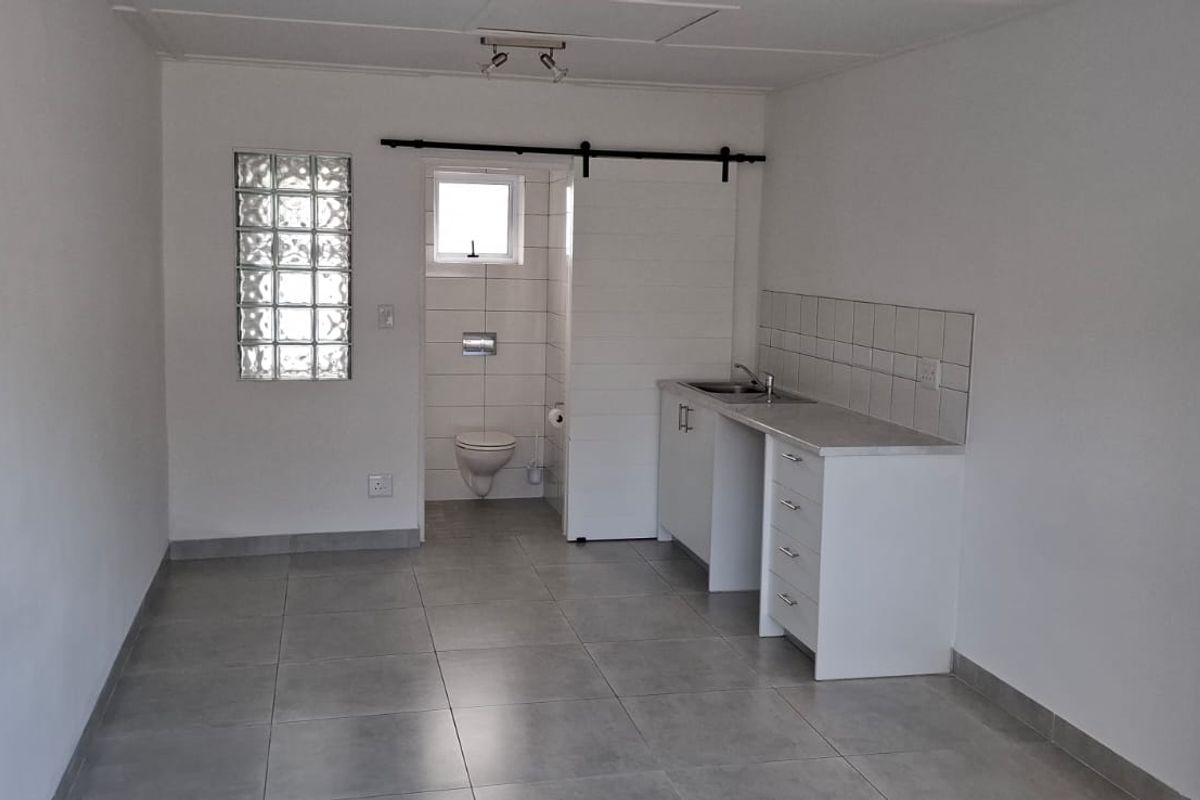 An open-plan studio space featuring tiled flooring, a compact kitchenette with sink, and an integrated bathroom with a wall-mounted toilet and glass block window.