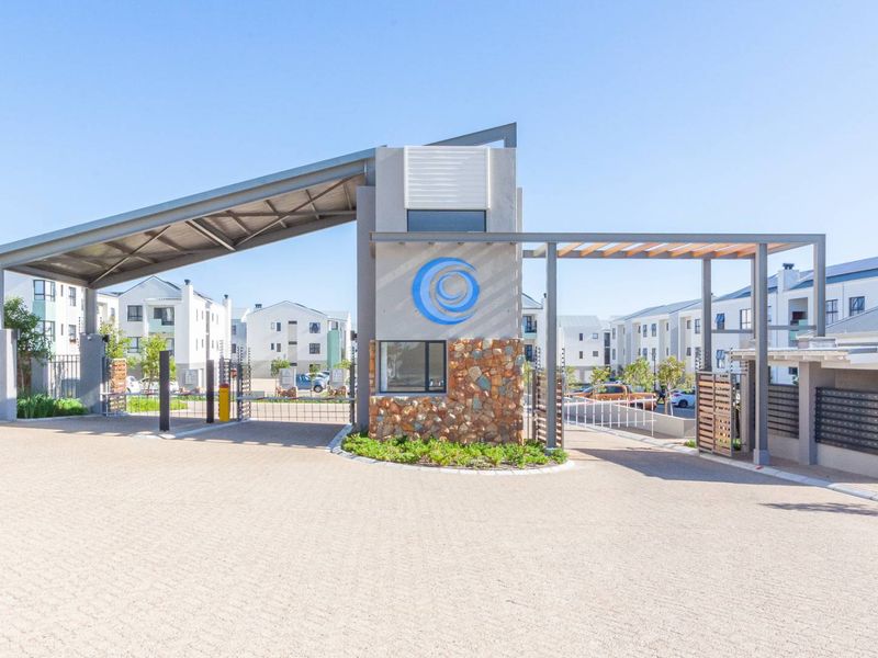 Modern apartment complex entrance featuring a secure gatehouse, stone-clad architectural details, and a paved driveway with automated access control.