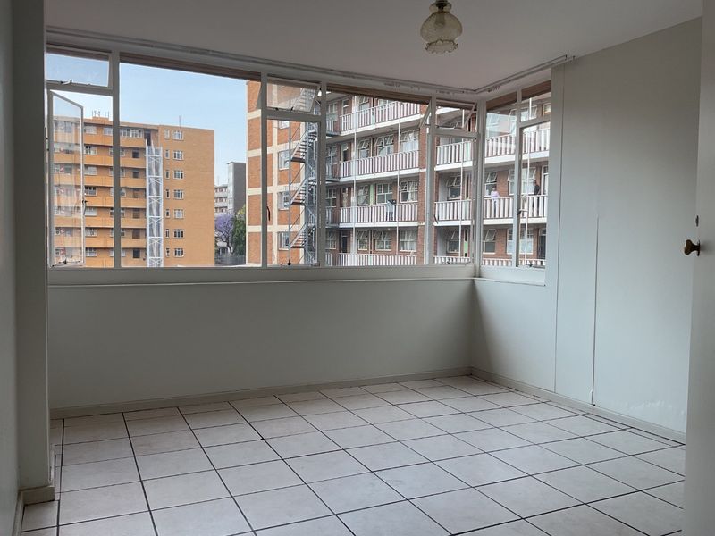 An empty, sunlit living area featuring tiled flooring and expansive wrap-around windows offering an urban view of the surrounding apartment complex.