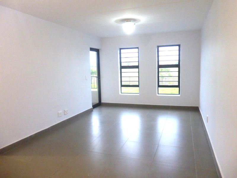 A bright, empty living area featuring neutral tiled flooring, large windows with security bars, and access to an outdoor space.