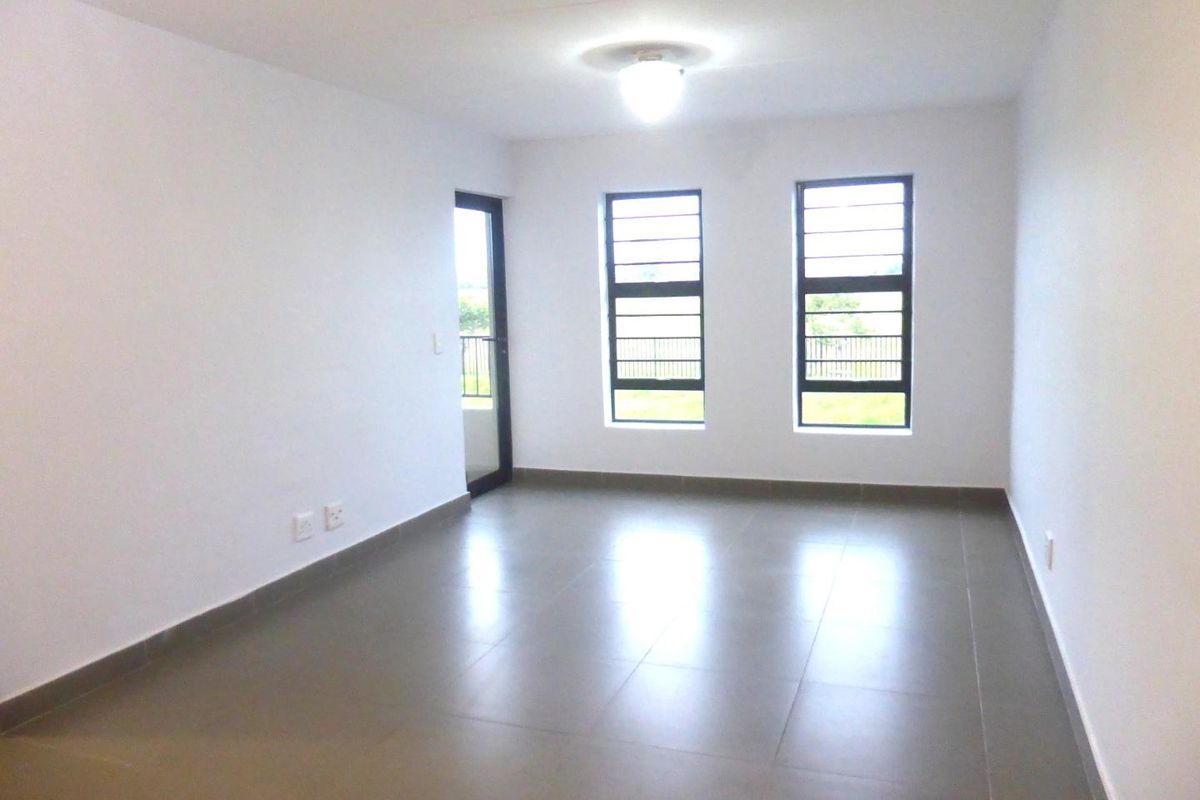 A bright, empty living area featuring neutral tiled flooring, large windows with security bars, and access to an outdoor space.