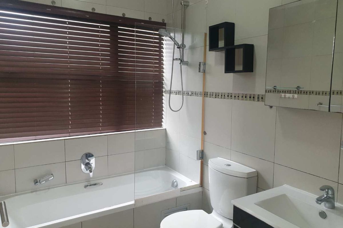A functional bathroom featuring a built-in bathtub with shower mixer, glass screen, wall-mounted toilet, and a vanity basin with mirror cabinet.