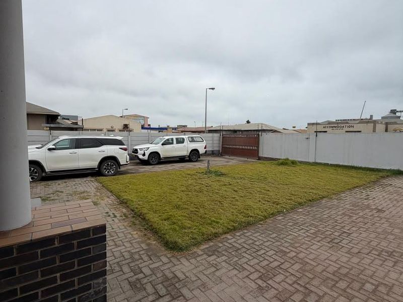 A view of the rear exterior area featuring a paved driveway, a small lawn, and a perimeter wall with a sliding gate.