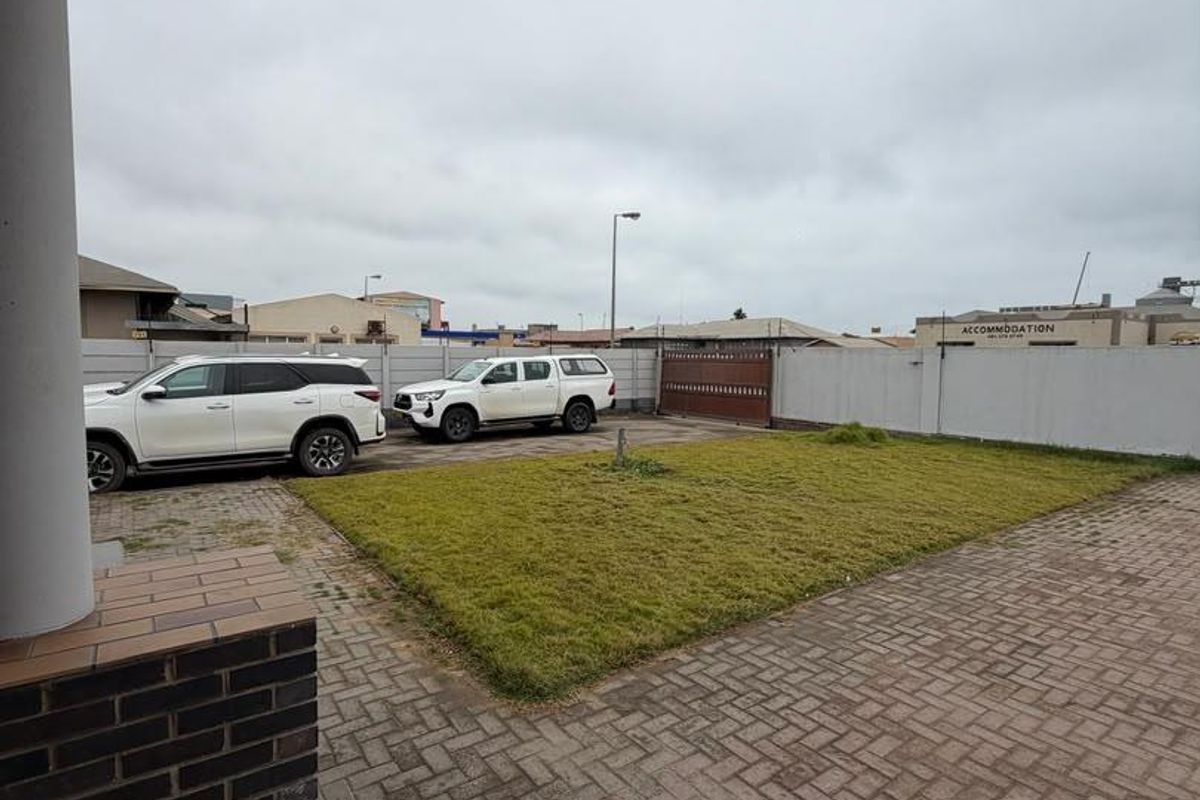 A view of the rear exterior area featuring a paved driveway, a small lawn, and a perimeter wall with a sliding gate.