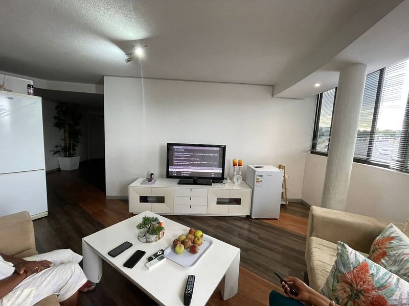 A living room featuring laminate flooring, a structural column, and large windows with venetian blinds, offering a functional and open living space.