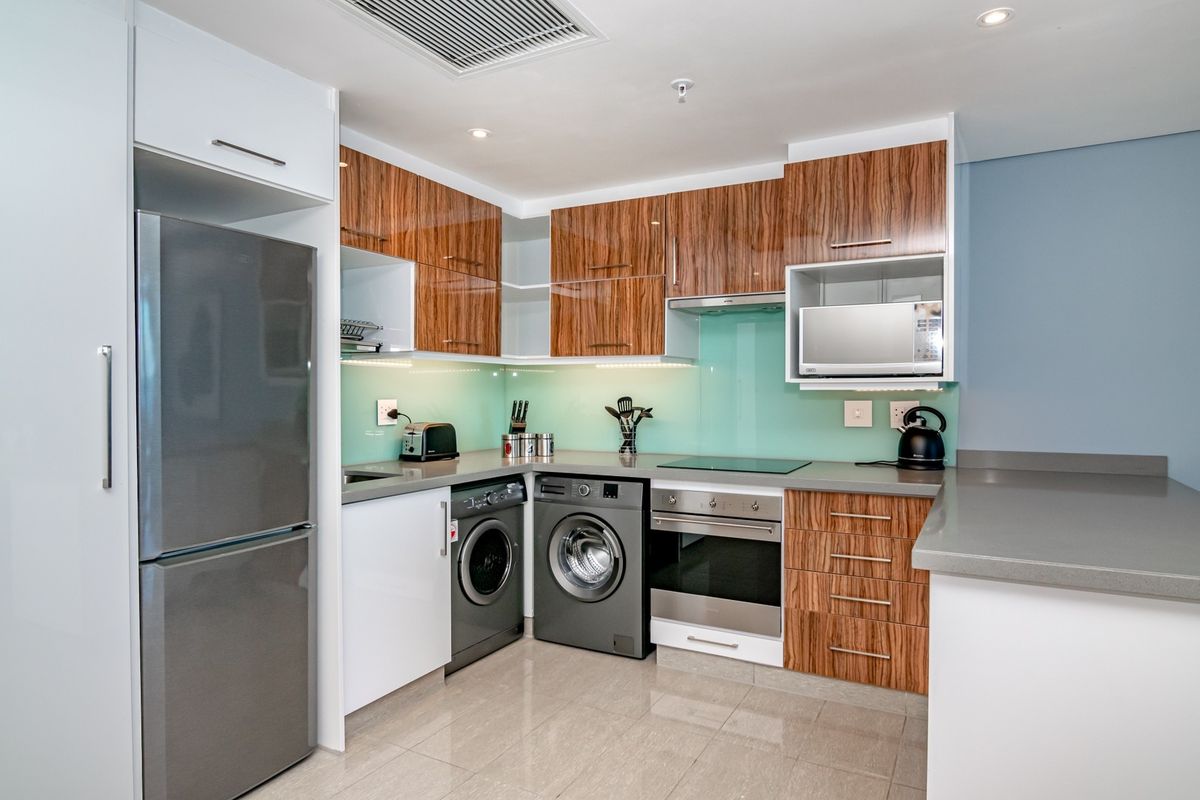 Modern kitchen featuring wood-grain cabinetry, stone countertops, a glass splashback, built-in oven, hob, and integrated laundry appliances.