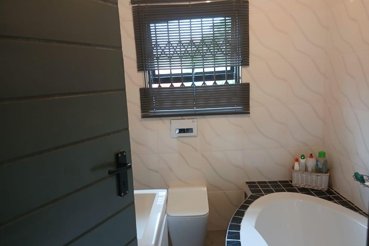 A compact bathroom featuring a built-in corner bathtub with tiled surround, a wall-mounted toilet, and a window with fitted blinds.