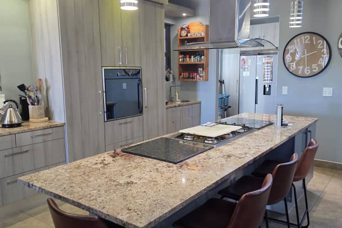 Modern kitchen featuring a large granite-topped island with integrated hob, stainless steel extractor fan, built-in oven, and contemporary cabinetry with recessed and pendant lighting.