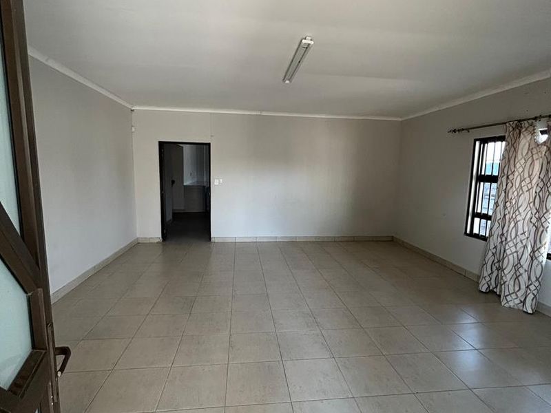 A spacious, empty living area featuring neutral ceramic floor tiles, clean walls, and a window with security bars.