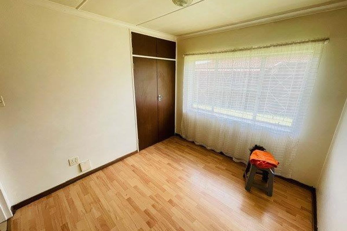 A bedroom featuring light wood-look flooring, a built-in cupboard with dark wood doors, and a large window with sheer curtains.