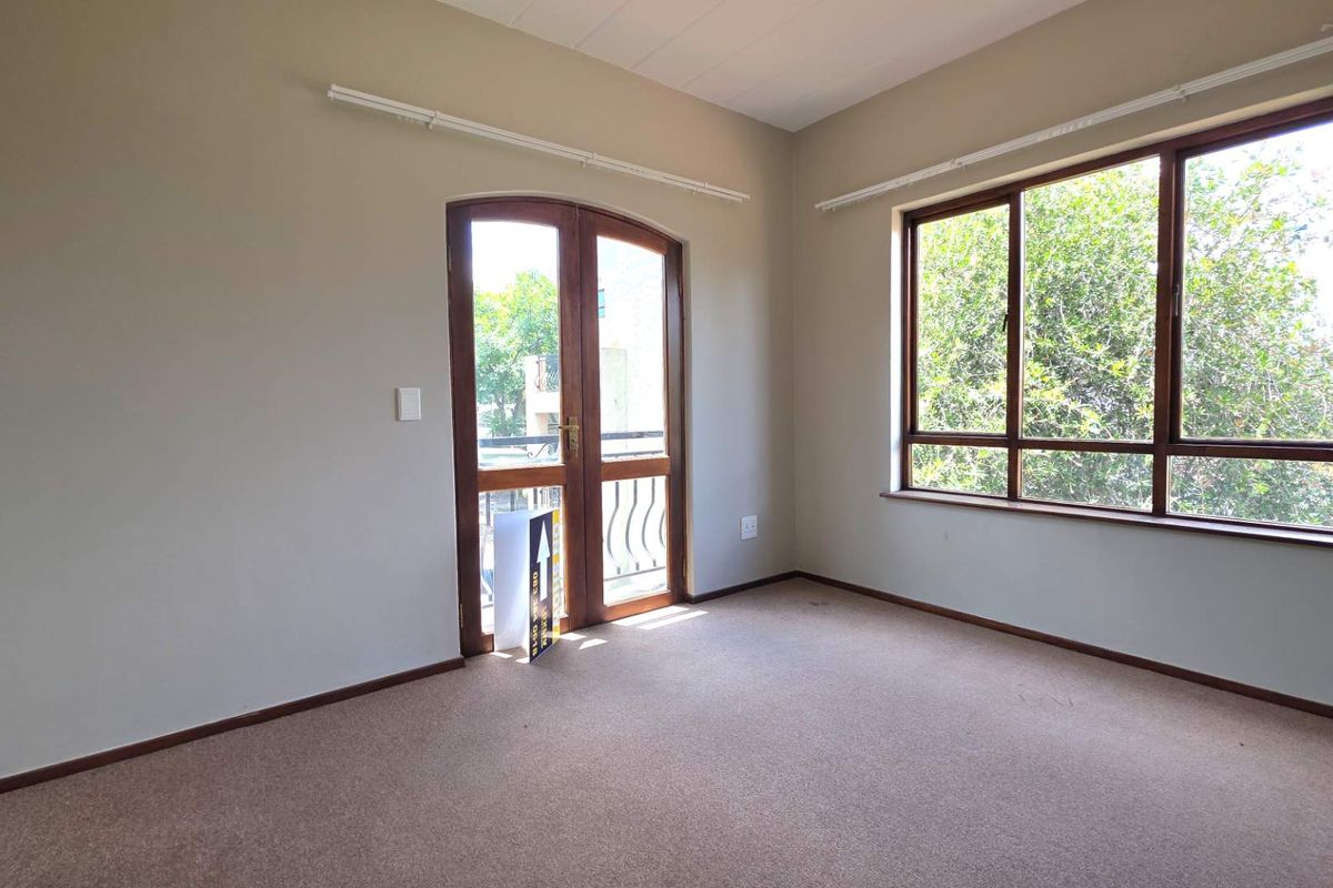 A bright, empty bedroom featuring neutral carpeting, large windows, and a glass-paneled door leading to a balcony.