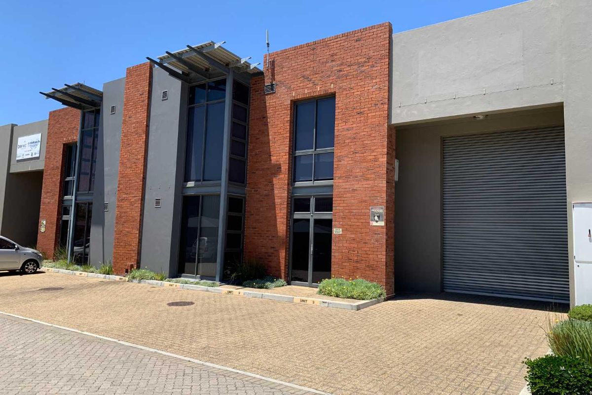 Modern industrial unit featuring a brick facade, large aluminium windows, and a secure roller shutter door with a paved driveway approach.