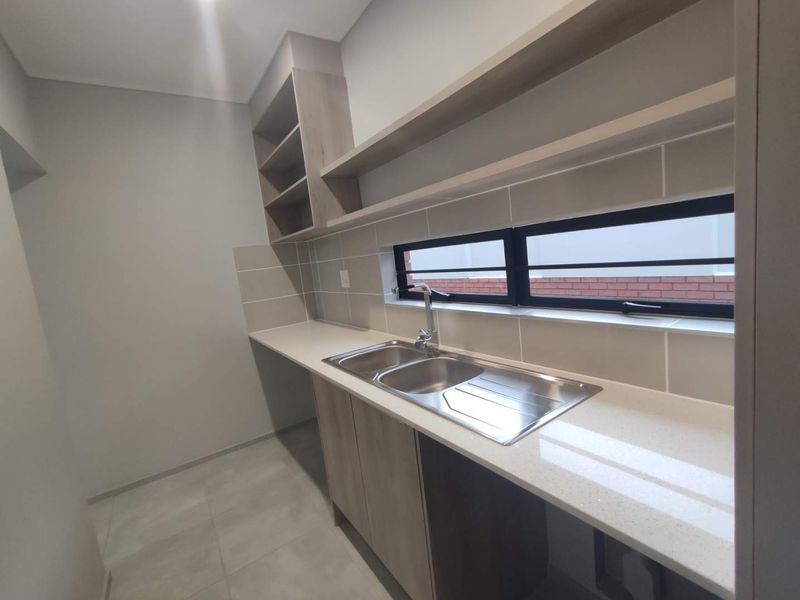A modern, clean scullery featuring stone countertops, double stainless steel sink, ample built-in cabinetry, and open shelving, finished with neutral wall tiles.