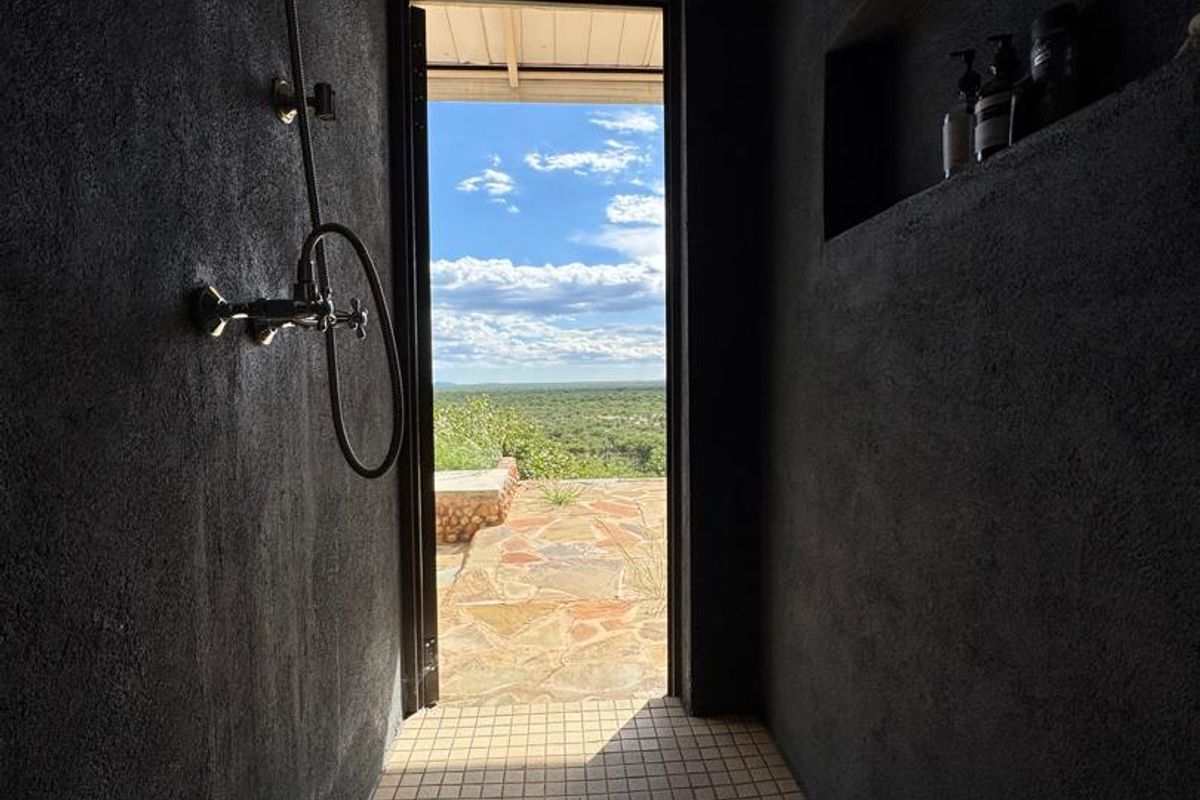 An open-air style shower featuring dark textured walls, tiled flooring, and a built-in wall niche, offering direct access to an outdoor patio with scenic views.