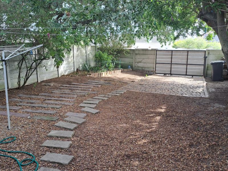A private rear garden area featuring a precast concrete boundary wall, sliding metal gate, stone paver pathway, and established trees.