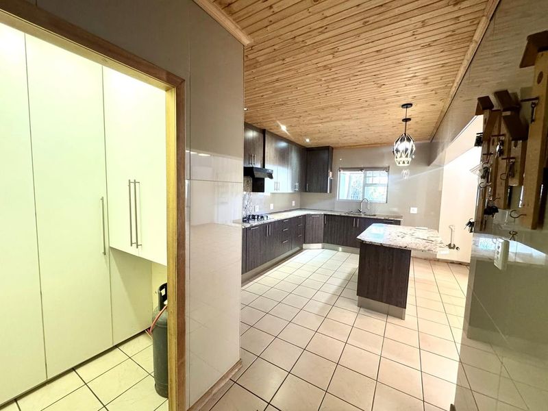 A spacious kitchen featuring dark wood cabinetry, stone countertops, a central island, tiled flooring, and a distinctive timber-clad ceiling.
