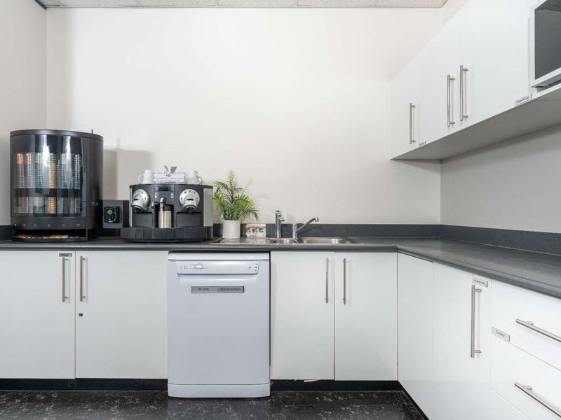 A functional office kitchenette featuring white cabinetry, dark laminate countertops, a stainless steel sink, and integrated appliances including a dishwasher and microwave.